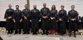 Pra todos verem: Nove pessoas estão alinhadas em pé, lado a lado, posando para foto. Todas veste...