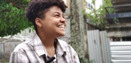Andy, pessoa negra, de perfil, sorrindo, vestida com camisa quadriculada.