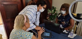 Imagem mostra uma idosa sendo atendida por duas profissionais, que usam máscara, em uma mesa com...