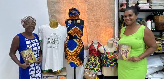 Imagem 02 mostra duas mulheres negras sorrindo. Uma senhora de 61 anos, que veste vestido azul e...