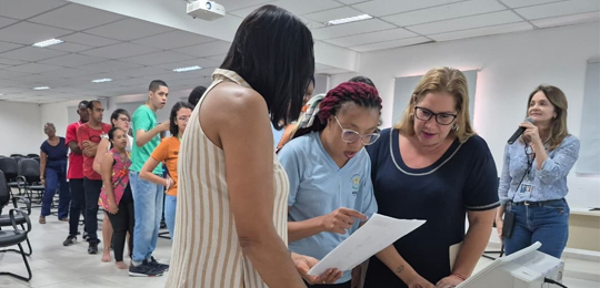 A imagem mostra pessoas reunidas em um espaço interno, semelhante a uma sala de eventos ou audit...