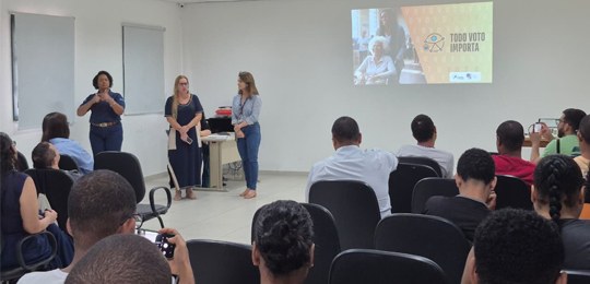 A imagem mostra um auditório com pessoas sentadas assistindo a uma apresentação. Três mulheres e...