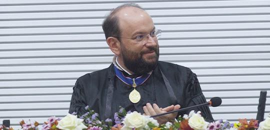 Pra todos verem: Homem, de pele clara, sentado em uma mesa, veste uma toga preta, com uma medalh...