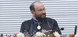 Pra todos verem: Homem, de pele clara, sentado em uma mesa, veste uma toga preta, com uma medalh...